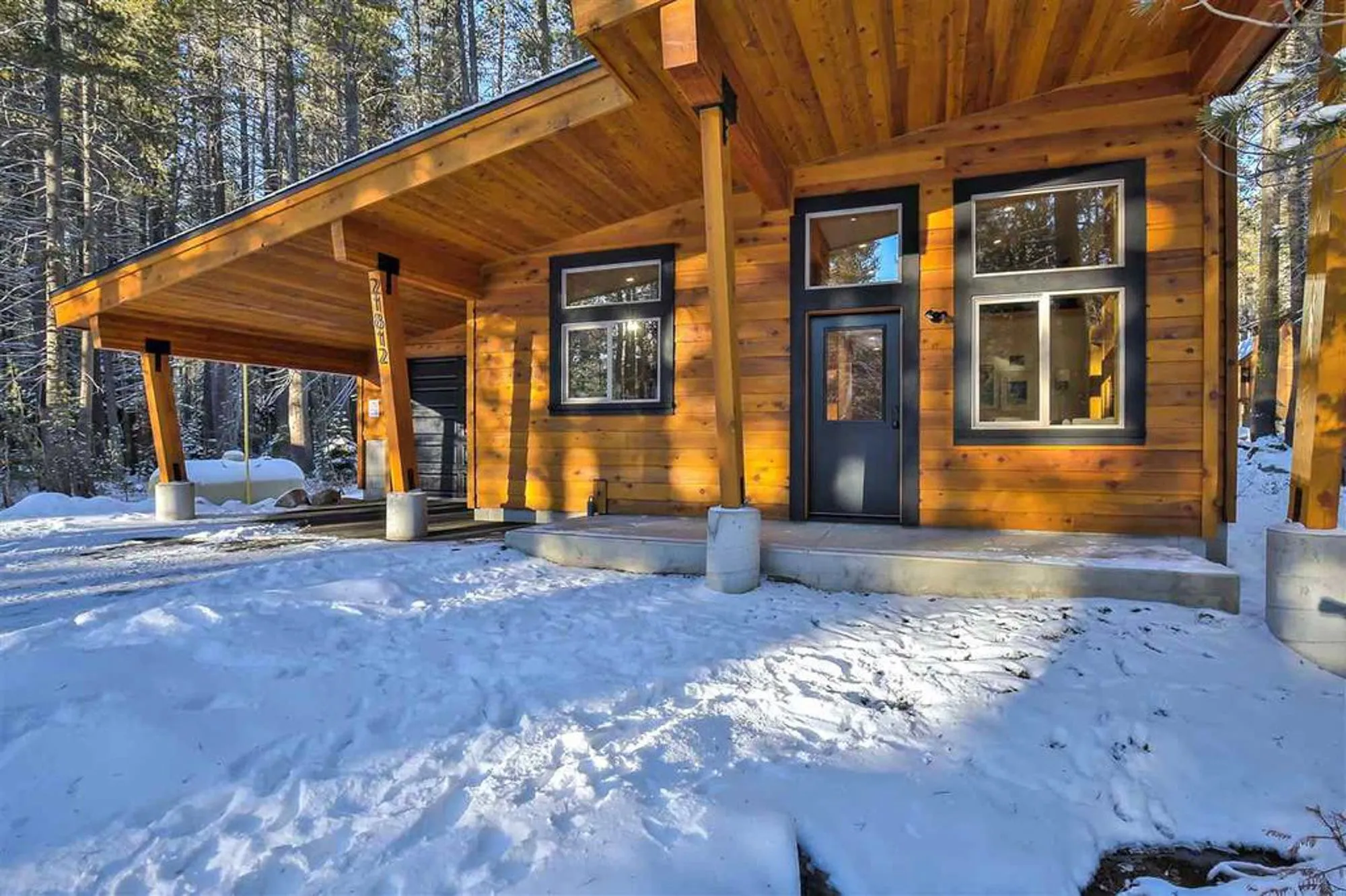 Small cedar-clad cabin with sloped roof, carport on timber posts and dark-framed windows in a snowy forest