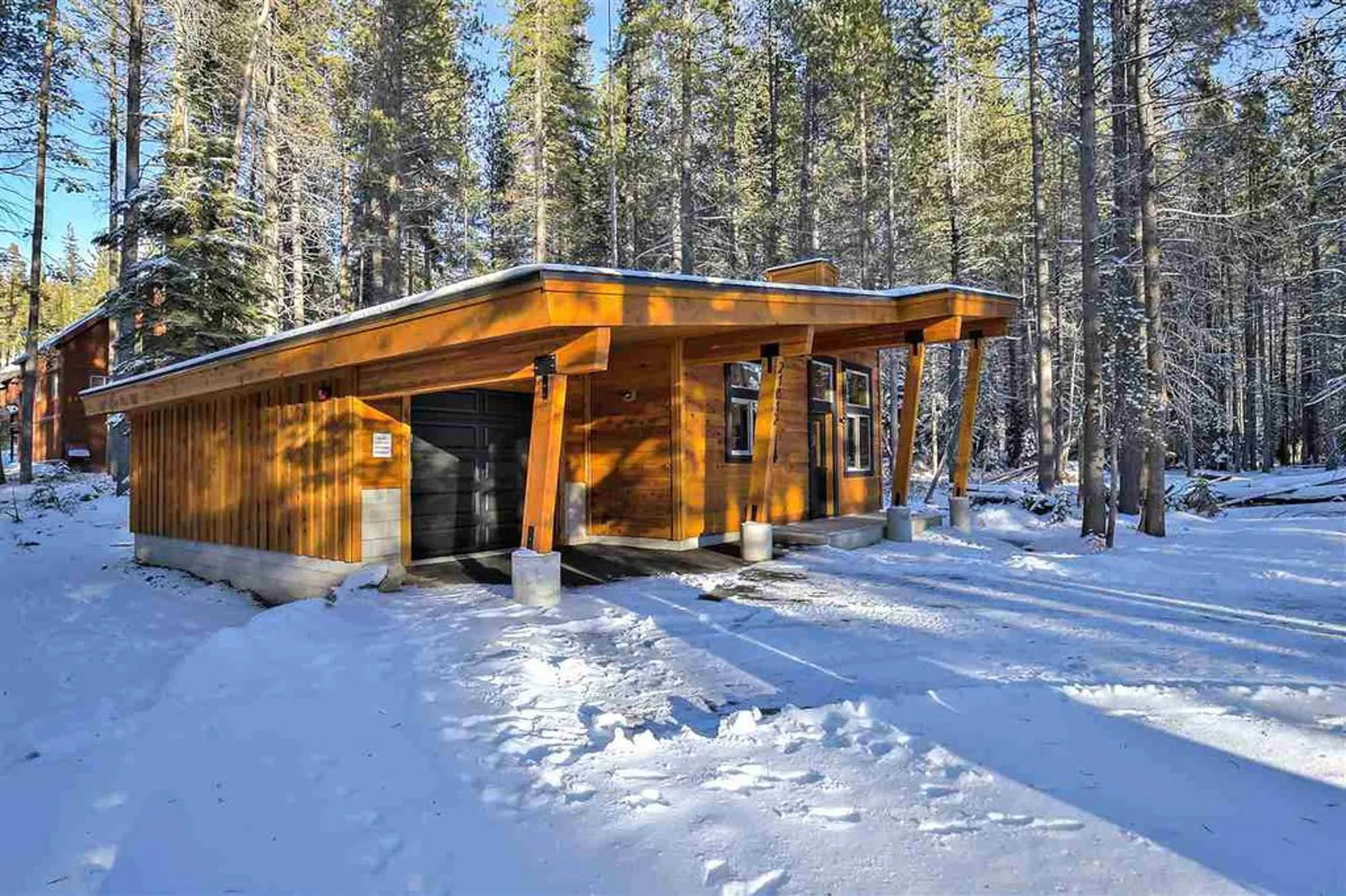 Side view of small wood-clad cabin with low shed roof, garage door and timber posts among snowy pines