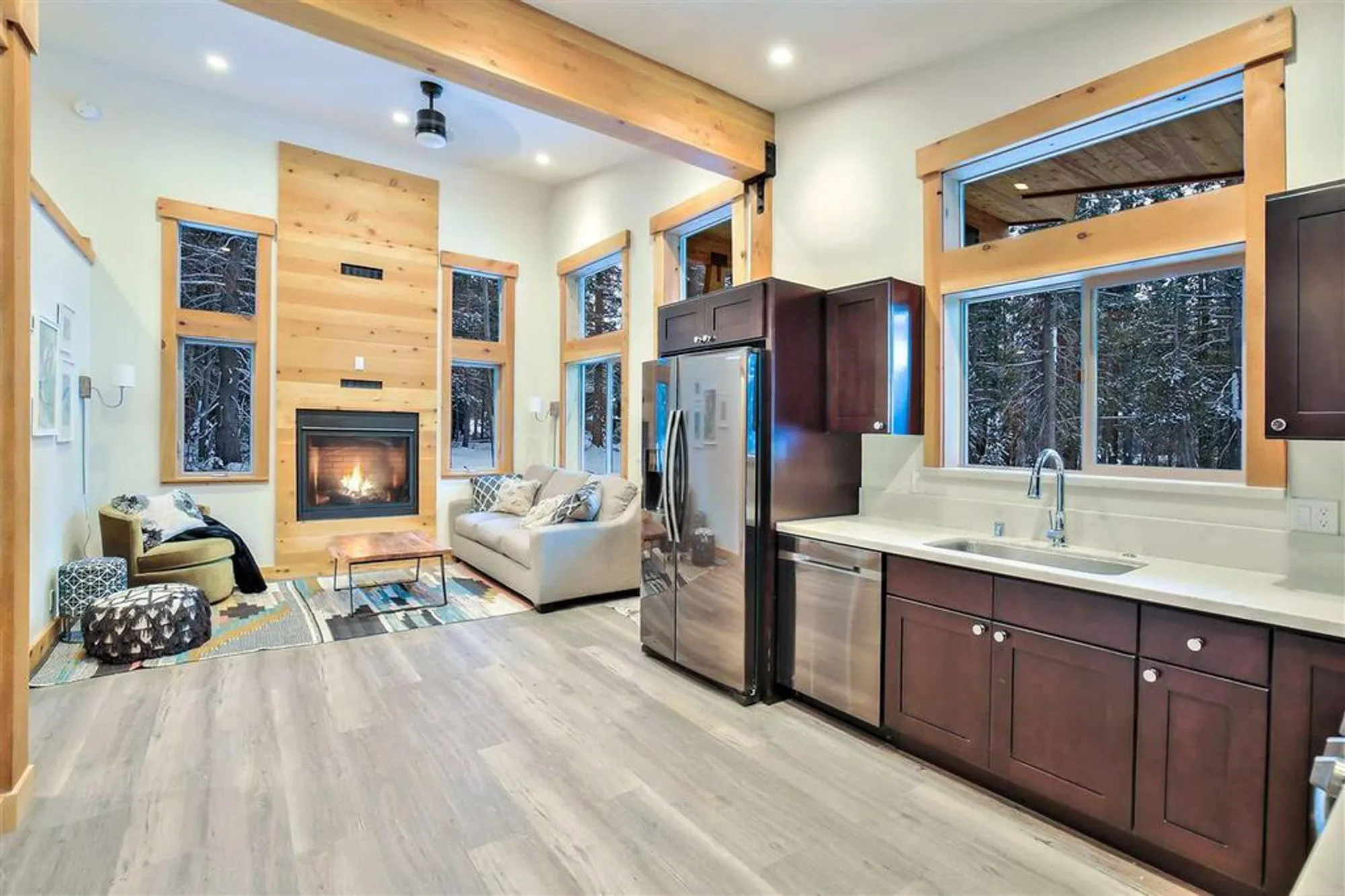 Tiny home interior with a wood-paneled fireplace wall, tall windows, sofa, and a compact kitchen with dark cabinets