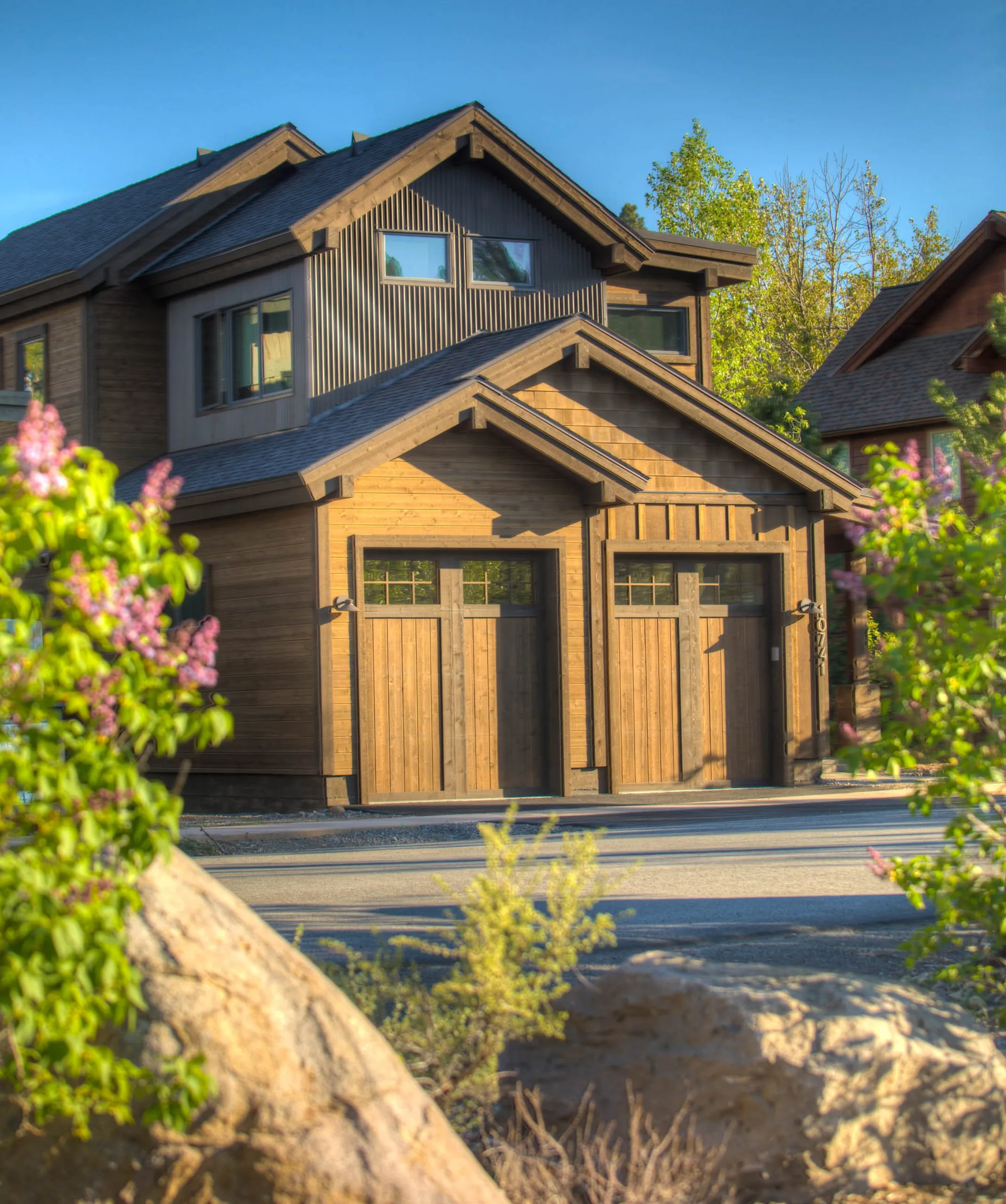 Home front with two wood garage doors, timber-trimmed gable roofs and dark siding, framed by blooming lilacs