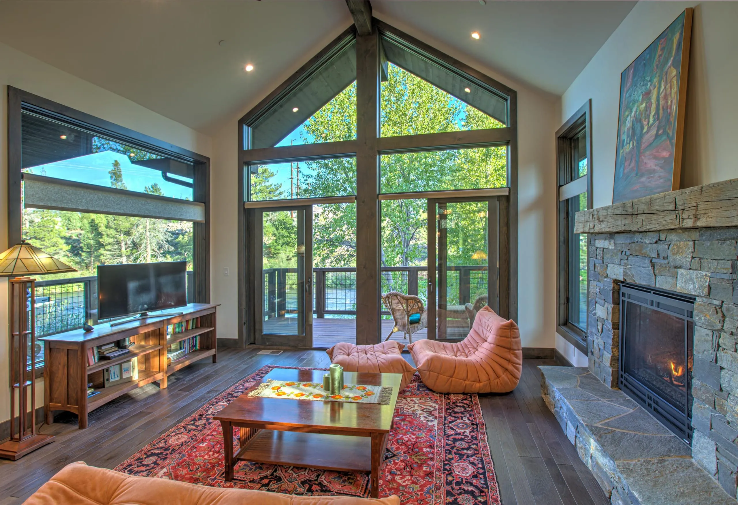 Living room with tall gabled window wall opening to a deck, stone fireplace and orange lounge chairs