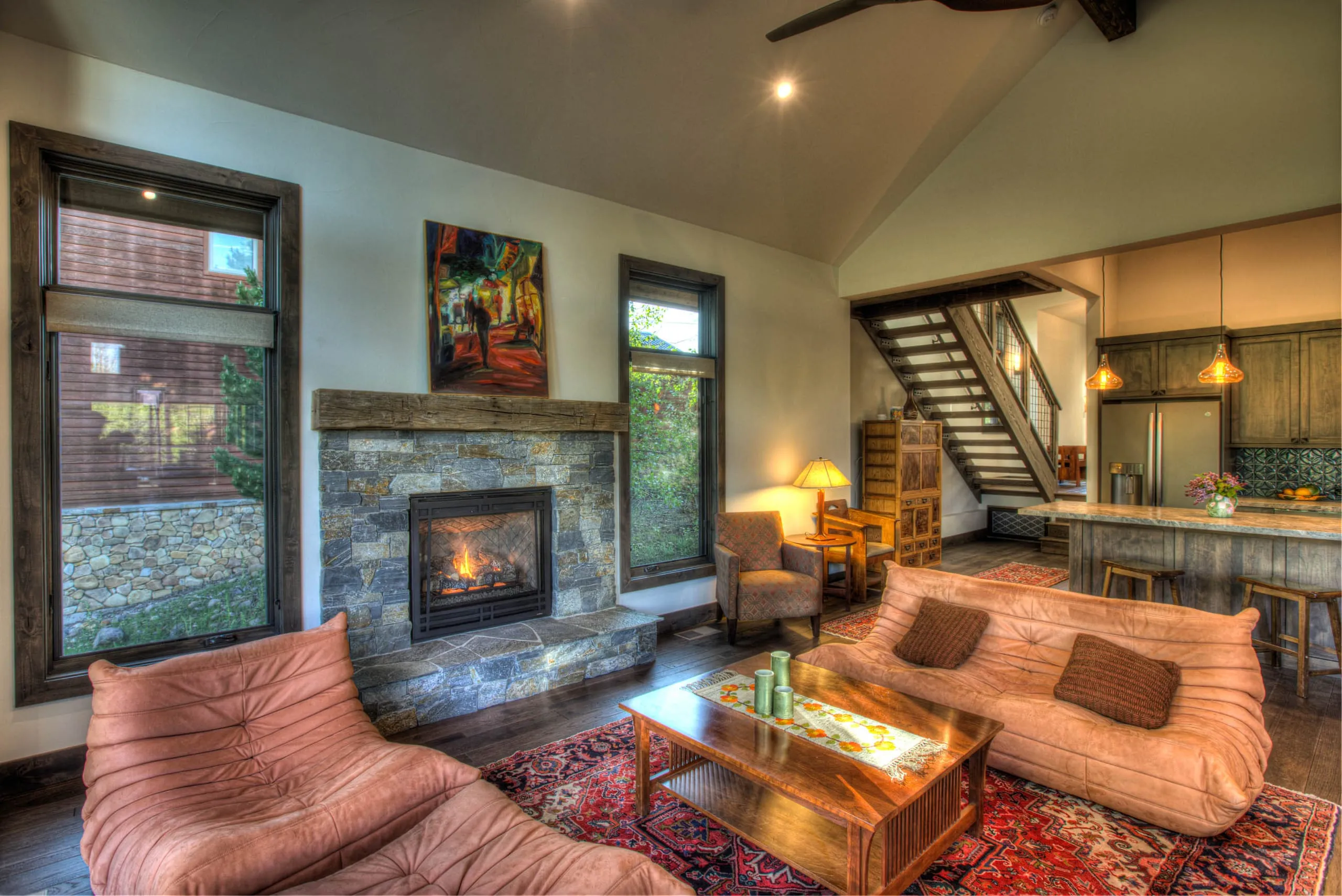 Living room with stone fireplace, large windows, orange sofa and chairs, and an open-tread staircase and kitchen beyond