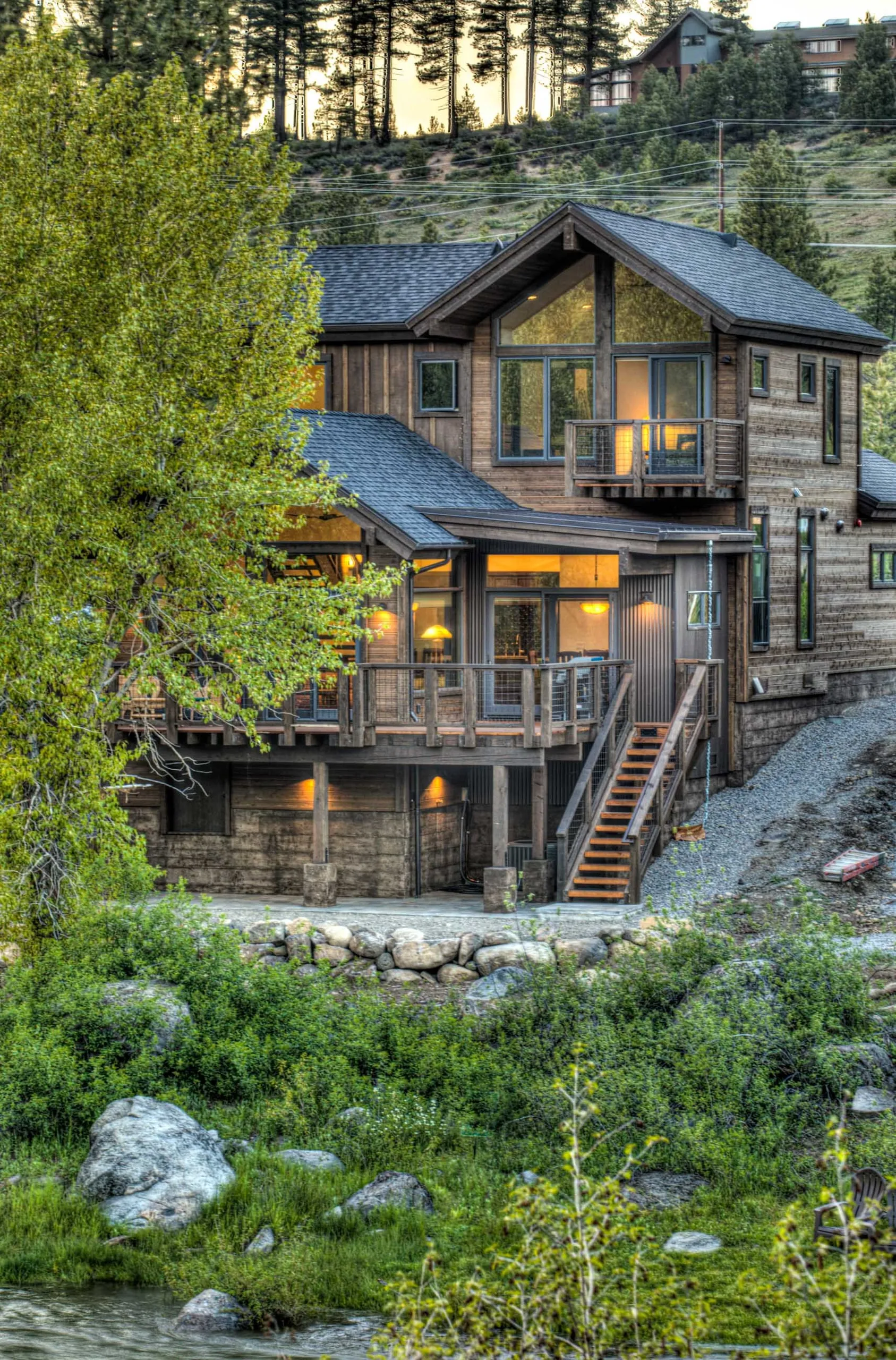 Riverside home with stone base, gabled roofs, balconies and exterior stair, lit at dusk above a green bank