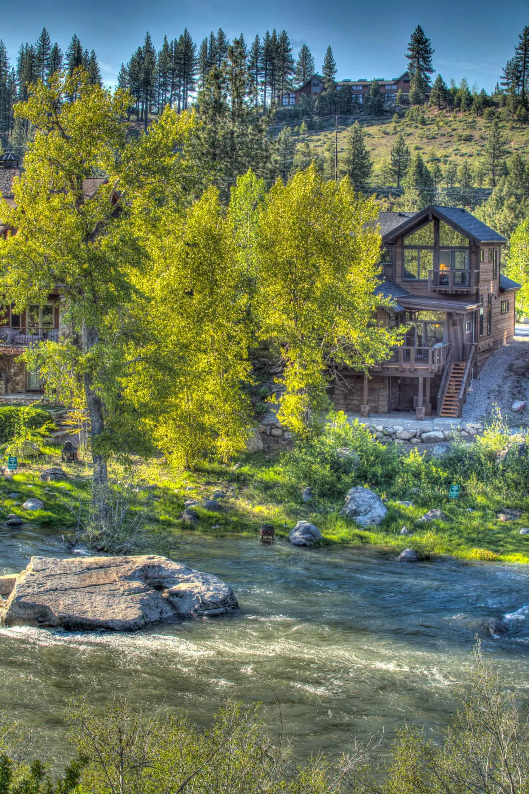 Rushing river with a large boulder in the foreground and a wood-sided home with decks on the bank