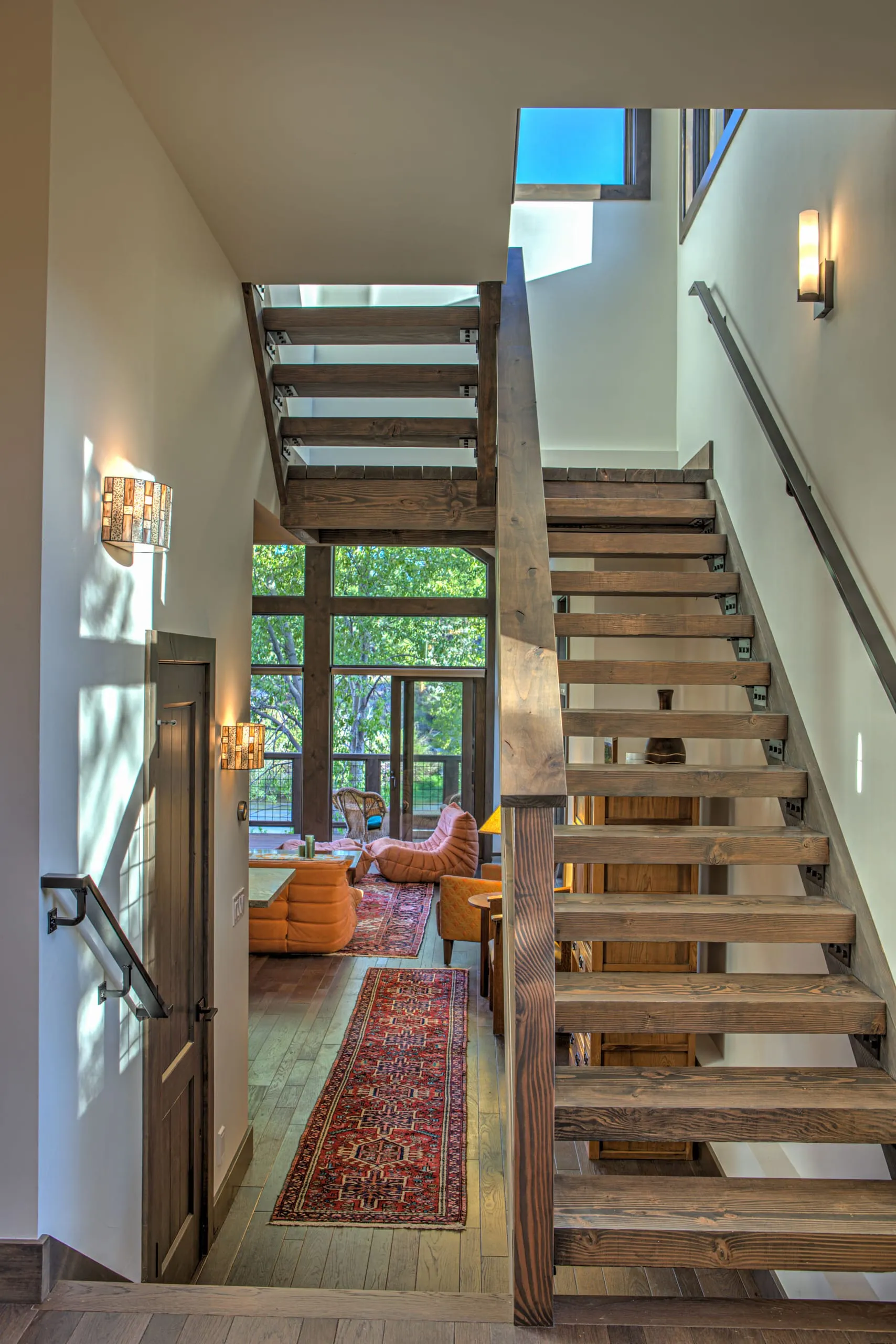 Open-tread timber staircase with a view down to a living room with tall windows and orange chairs