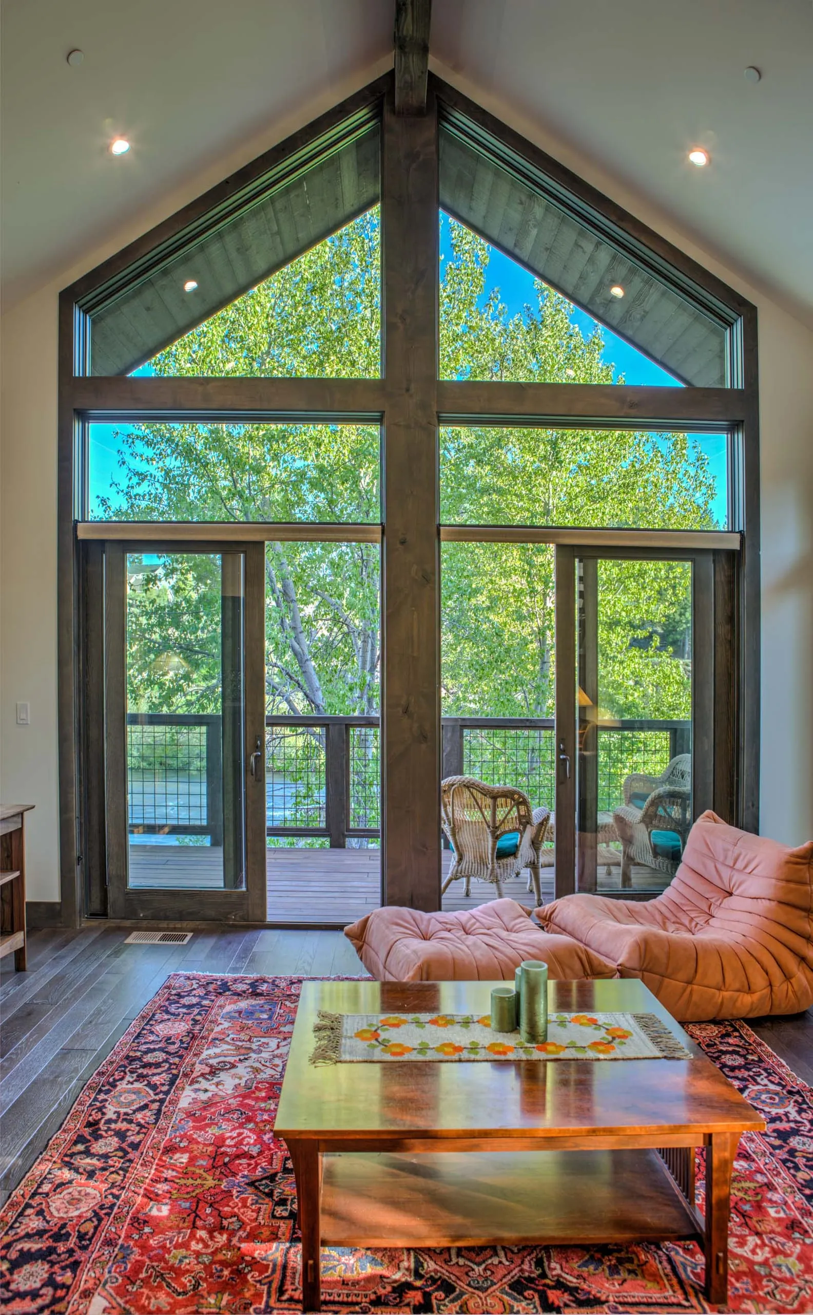 Tall living room window wall with timber frame, glass doors to a deck, orange chairs and red rug