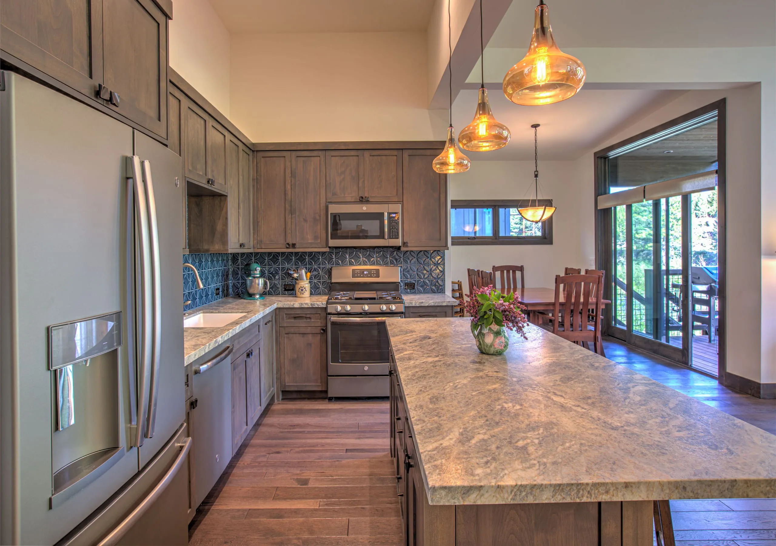 Kitchen with gray-brown cabinets, blue patterned tile backsplash, stone-topped island, pendant lights and dining table beyond