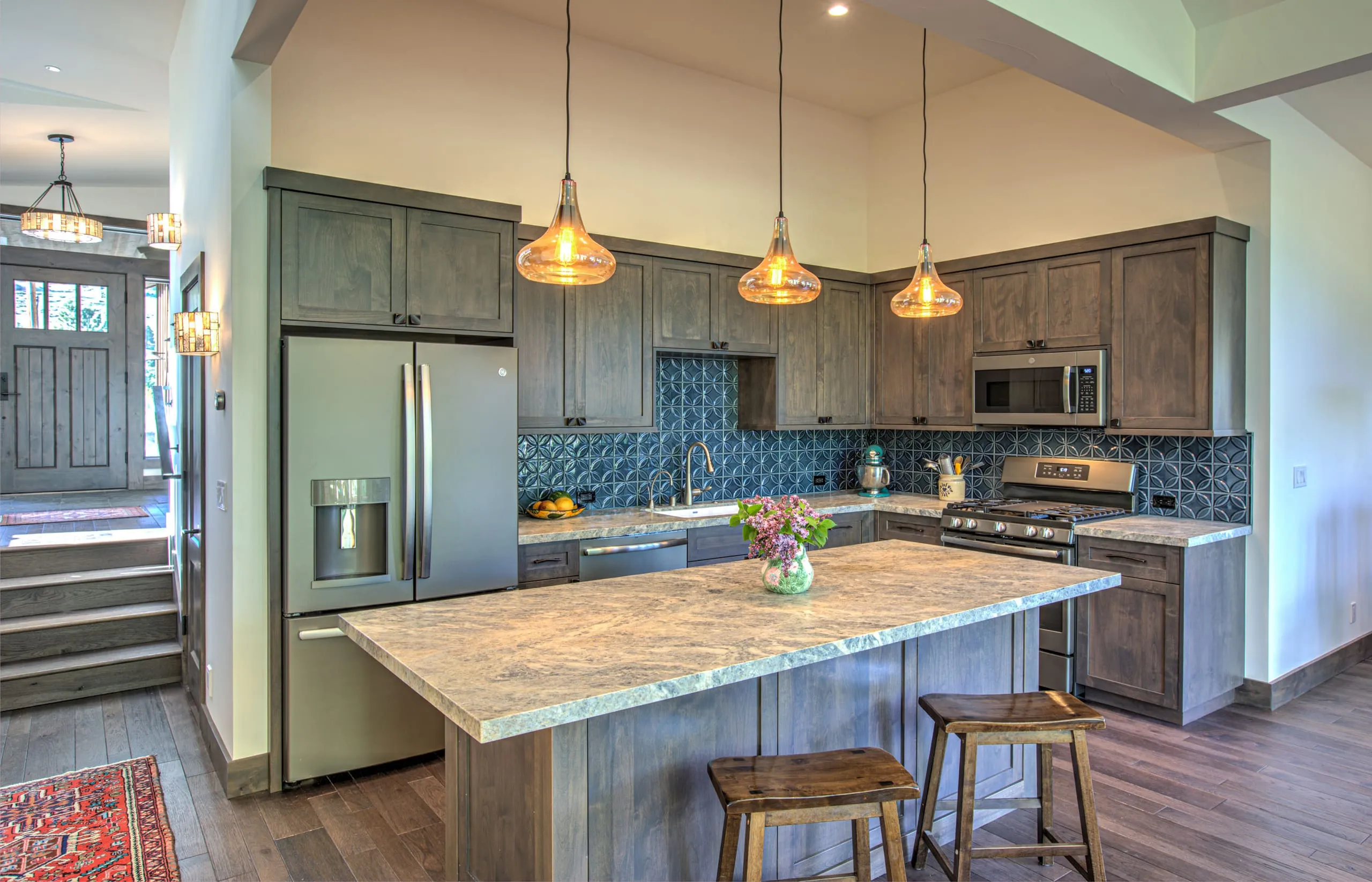 Kitchen with blue tile backsplash, stone island with wood stools, pendant lights and an entry stair beyond