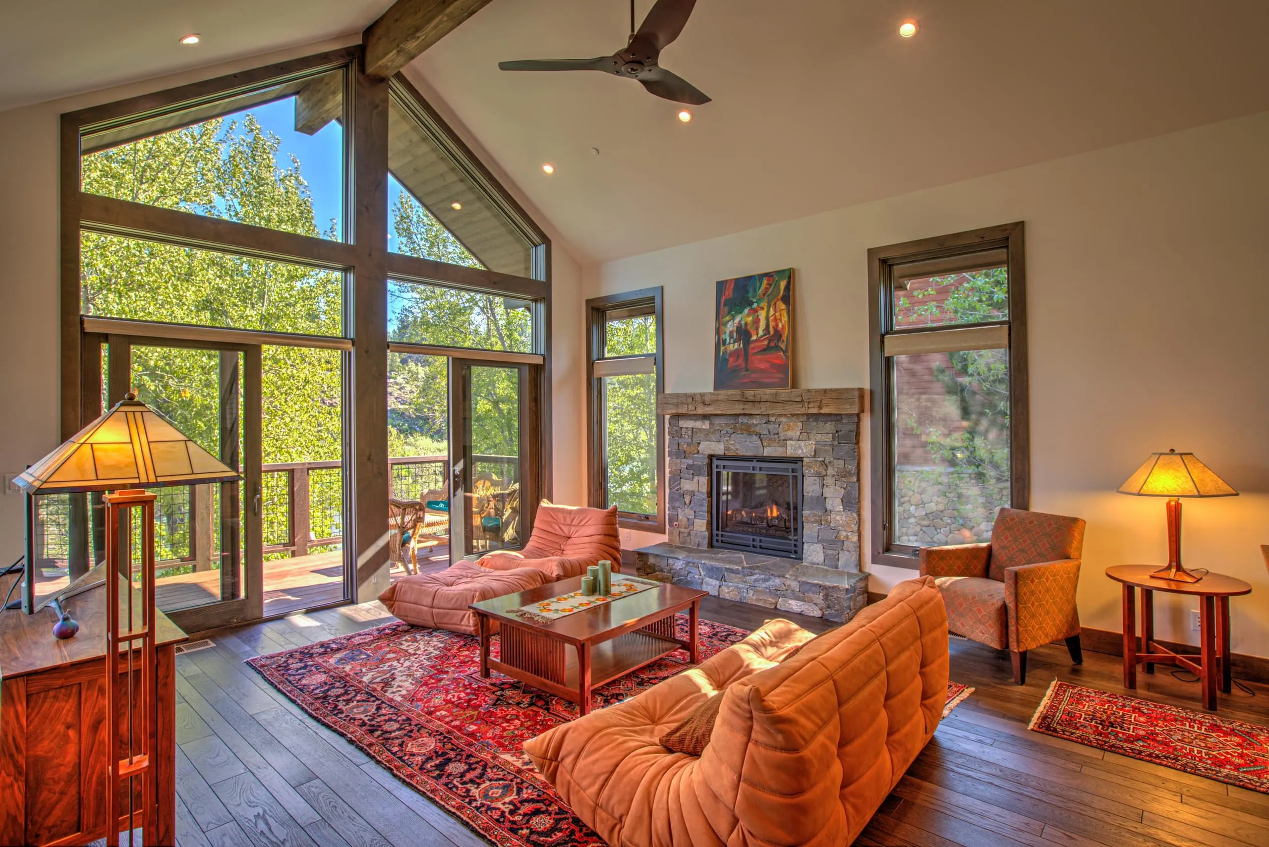 Vaulted living room with large corner windows, stone fireplace, orange sofa and armchair, and a red rug