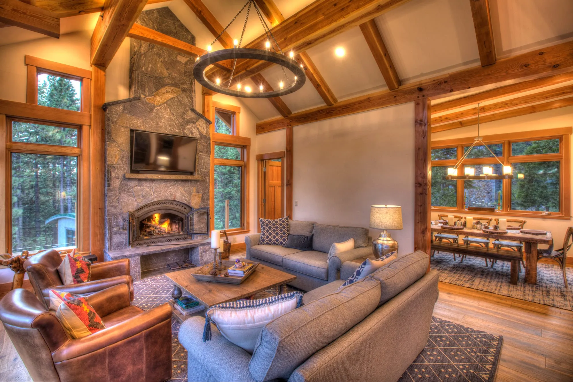 Timber-frame great room with stone fireplace, wood-framed windows, gray sofas, leather chairs and a dining table at right