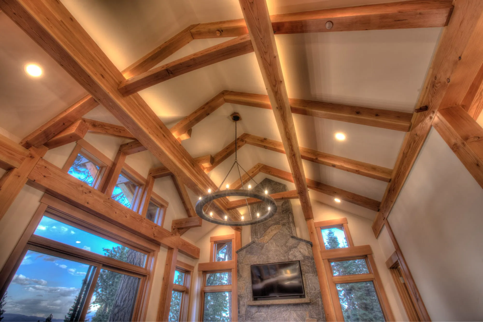 Upward view of exposed timber roof trusses, round iron chandelier and a stone fireplace chimney with large windows