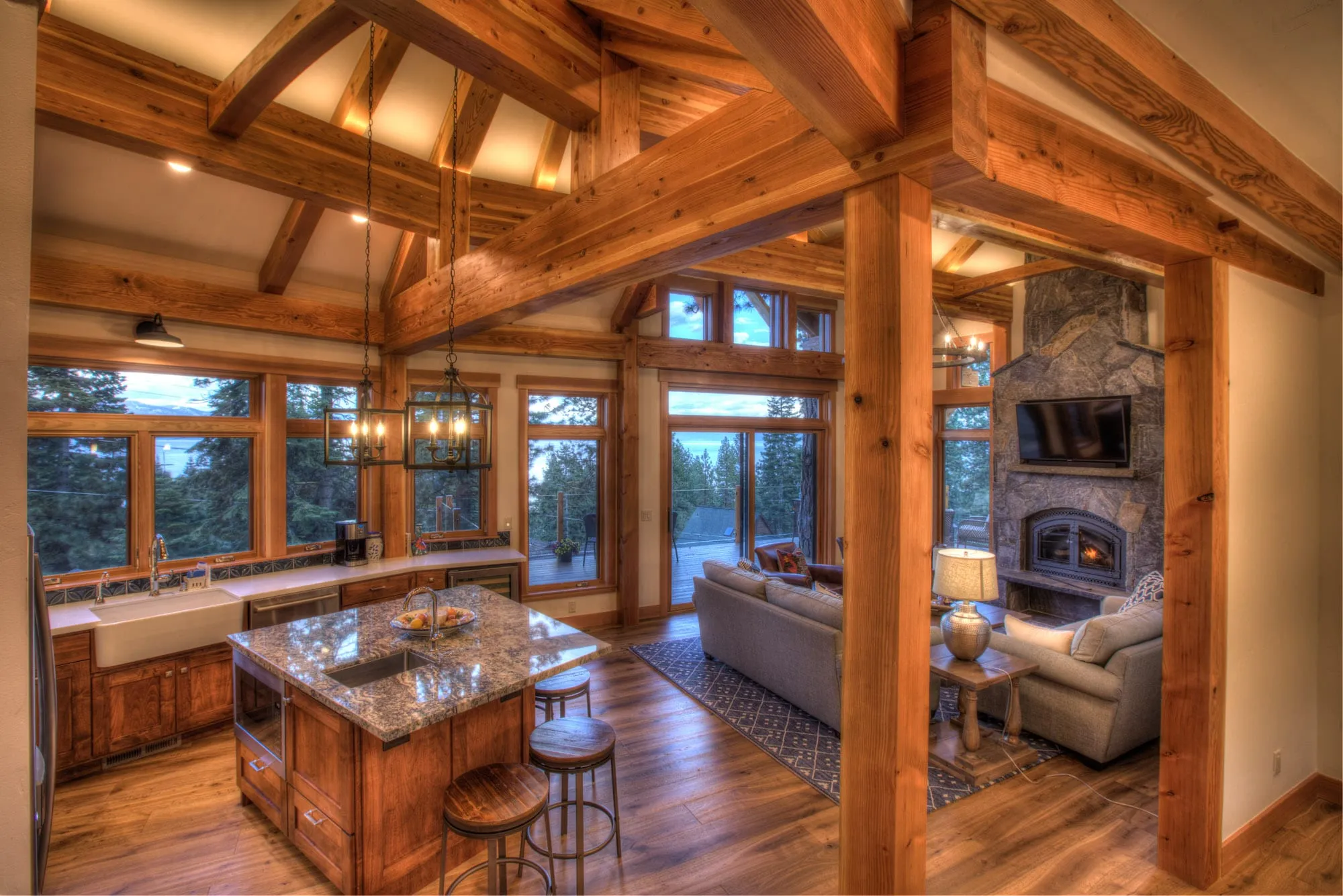 Open timber-frame kitchen and living area with granite island, wood posts, stone fireplace and forest views through windows