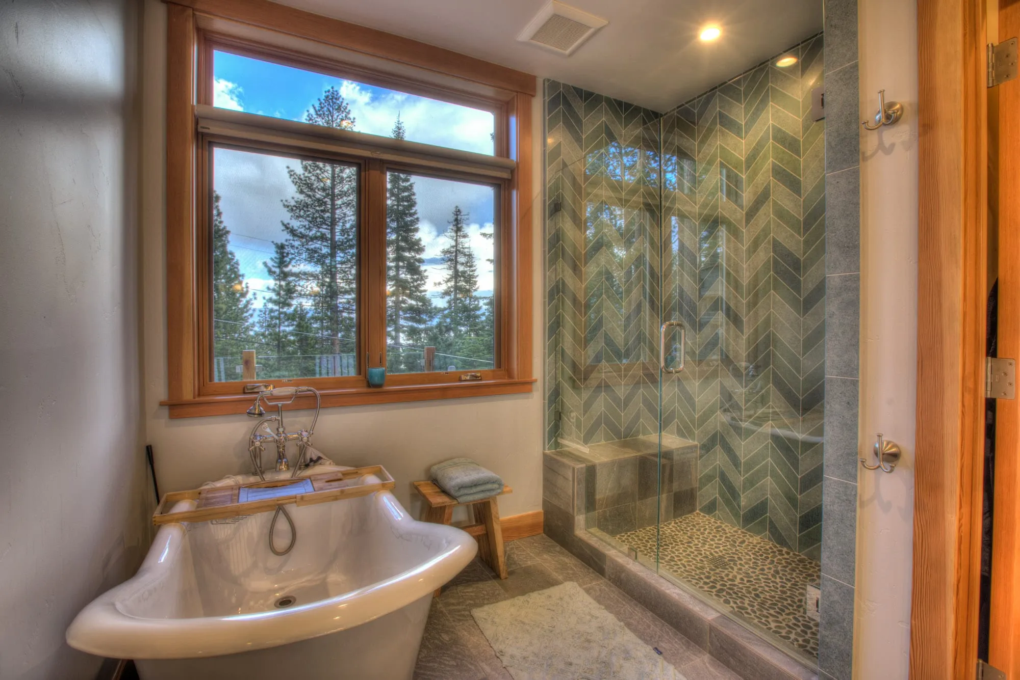 Bathroom with freestanding soaking tub, chevron-tiled glass shower and a wood-framed window facing pine trees