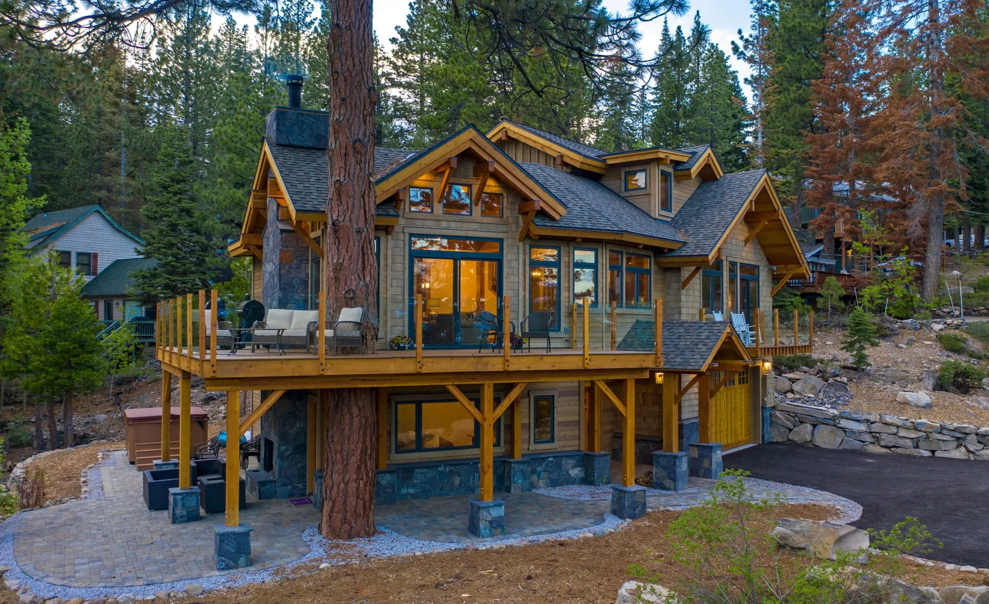 Timber-frame home exterior at dusk with wraparound deck built around a large pine trunk, stone base and paver patio