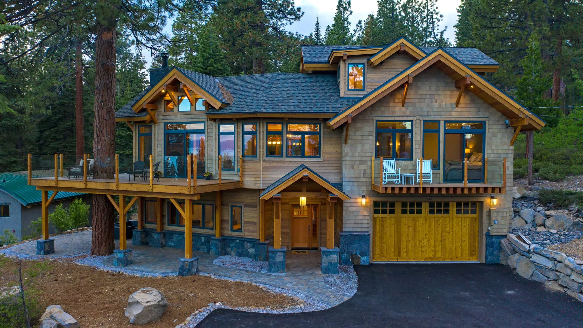 Front of a shingle-clad timber-frame home with gabled entry, yellow wood garage doors, upper deck and stone base