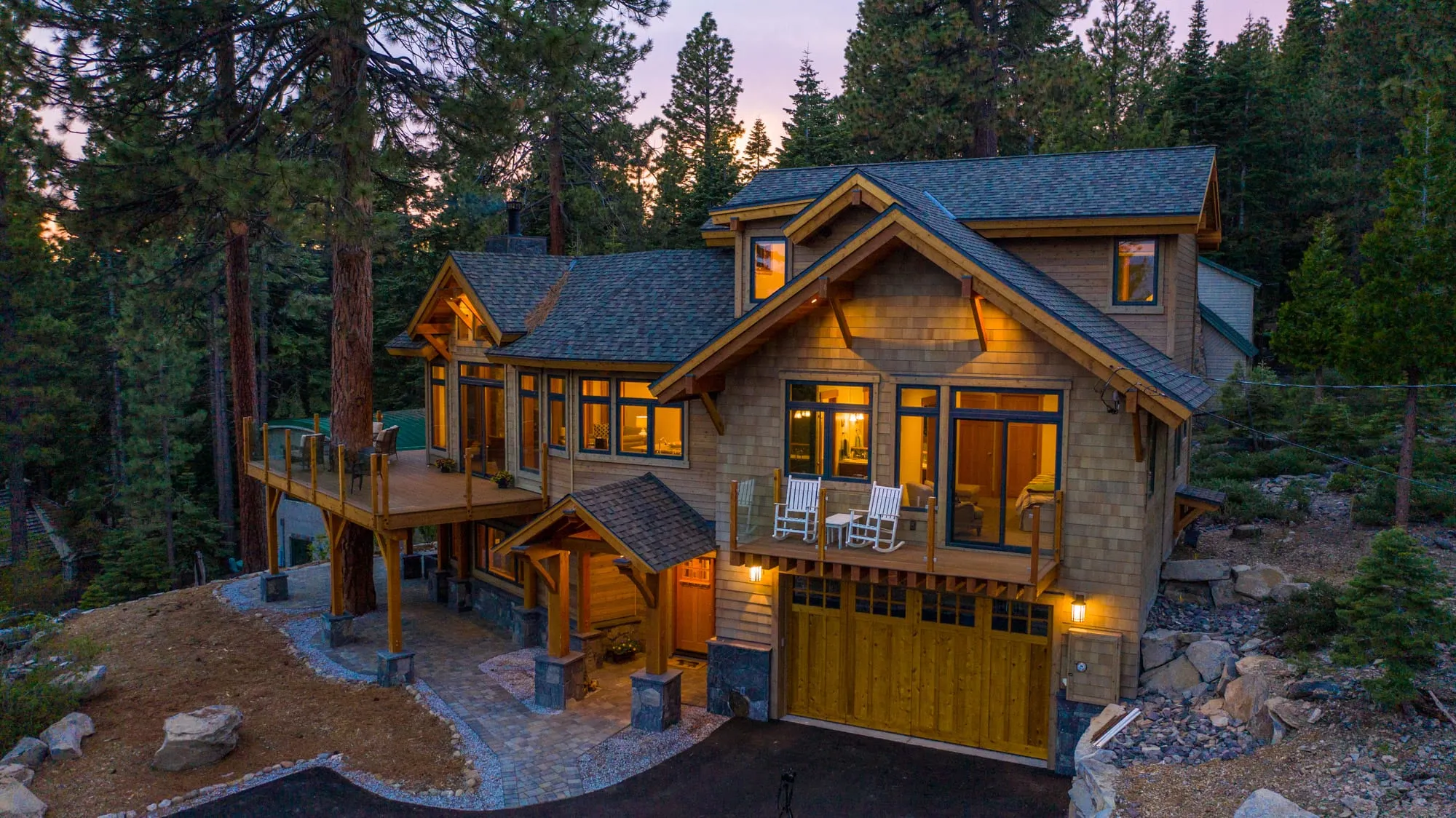 Aerial dusk view of a timber-frame mountain home with glowing windows, deck around a tree and wood garage doors