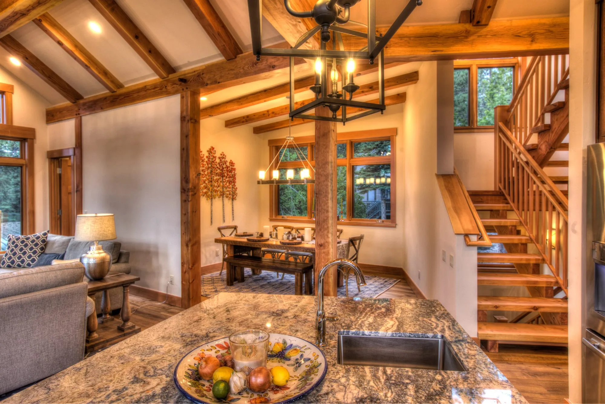 Open kitchen island with granite counter looking toward a timber-beamed dining area, living room and wood staircase