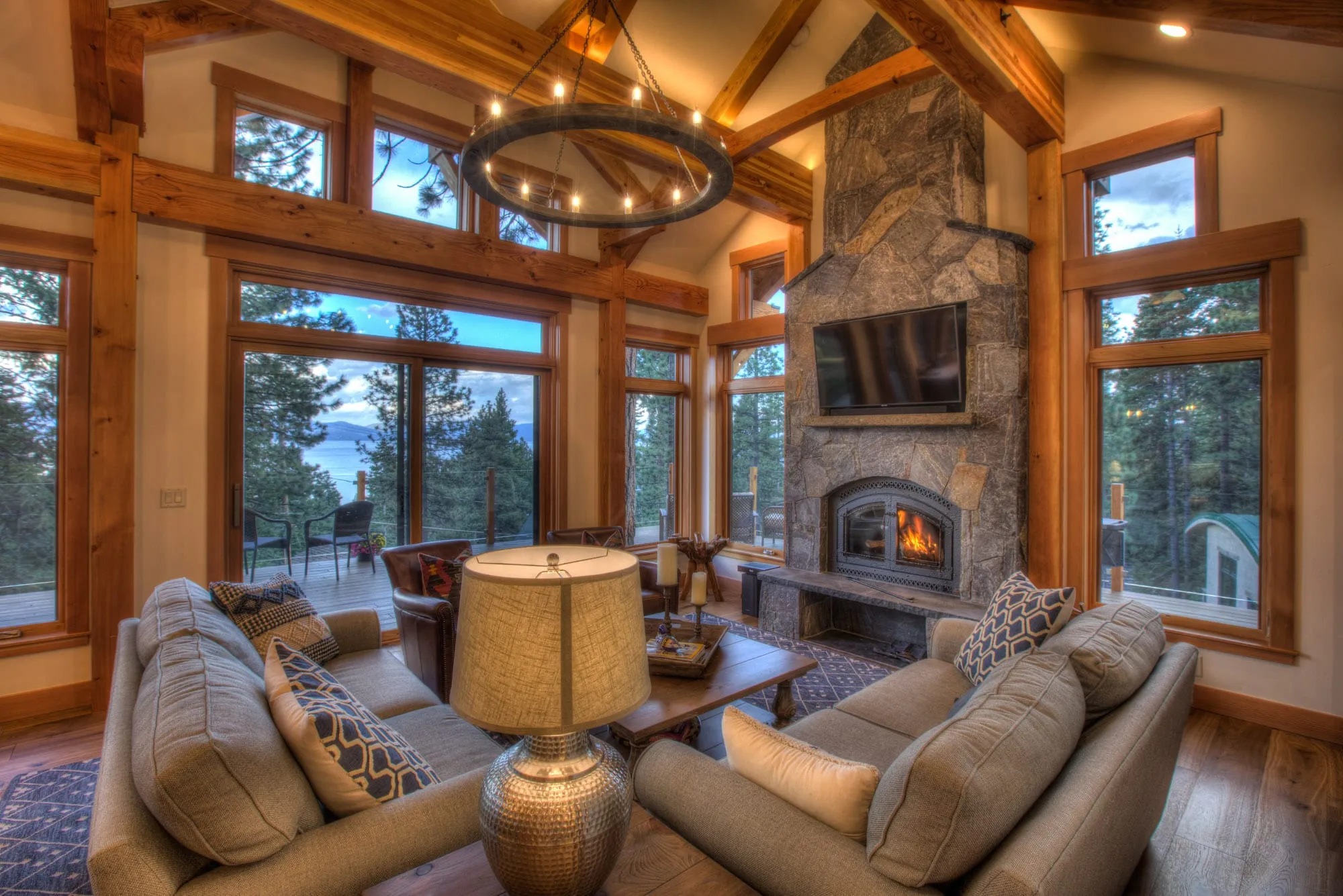 Timber-frame great room with stone fireplace, tall windows, wood floors and a distant lake view through the glass doors