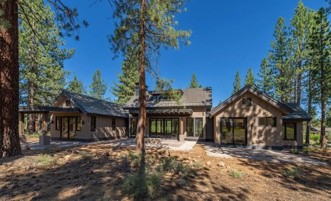 Rear of a single-story home with gabled wings, dark roofs and glass doors opening to a terrace among tall pines
