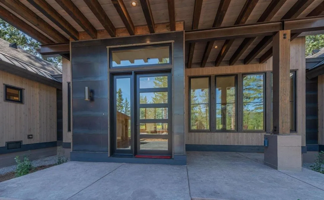 Covered entry under exposed wood rafters, with a dark metal-clad wall, a glass front door and tall windows