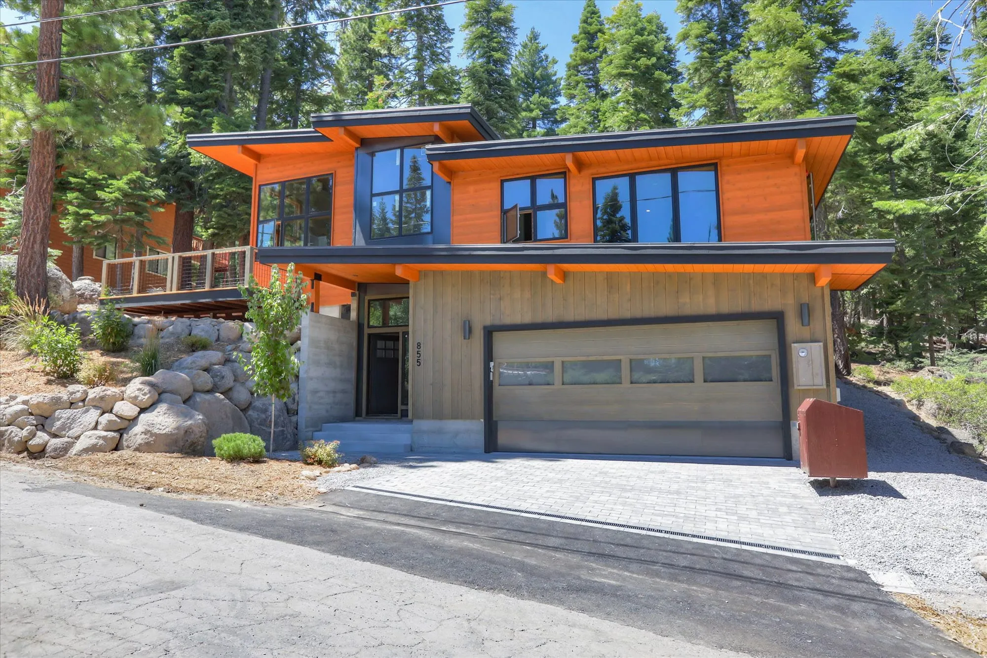 Street-side view of a contemporary home with shed roofs, orange wood siding, two-car garage and rock-lined slope