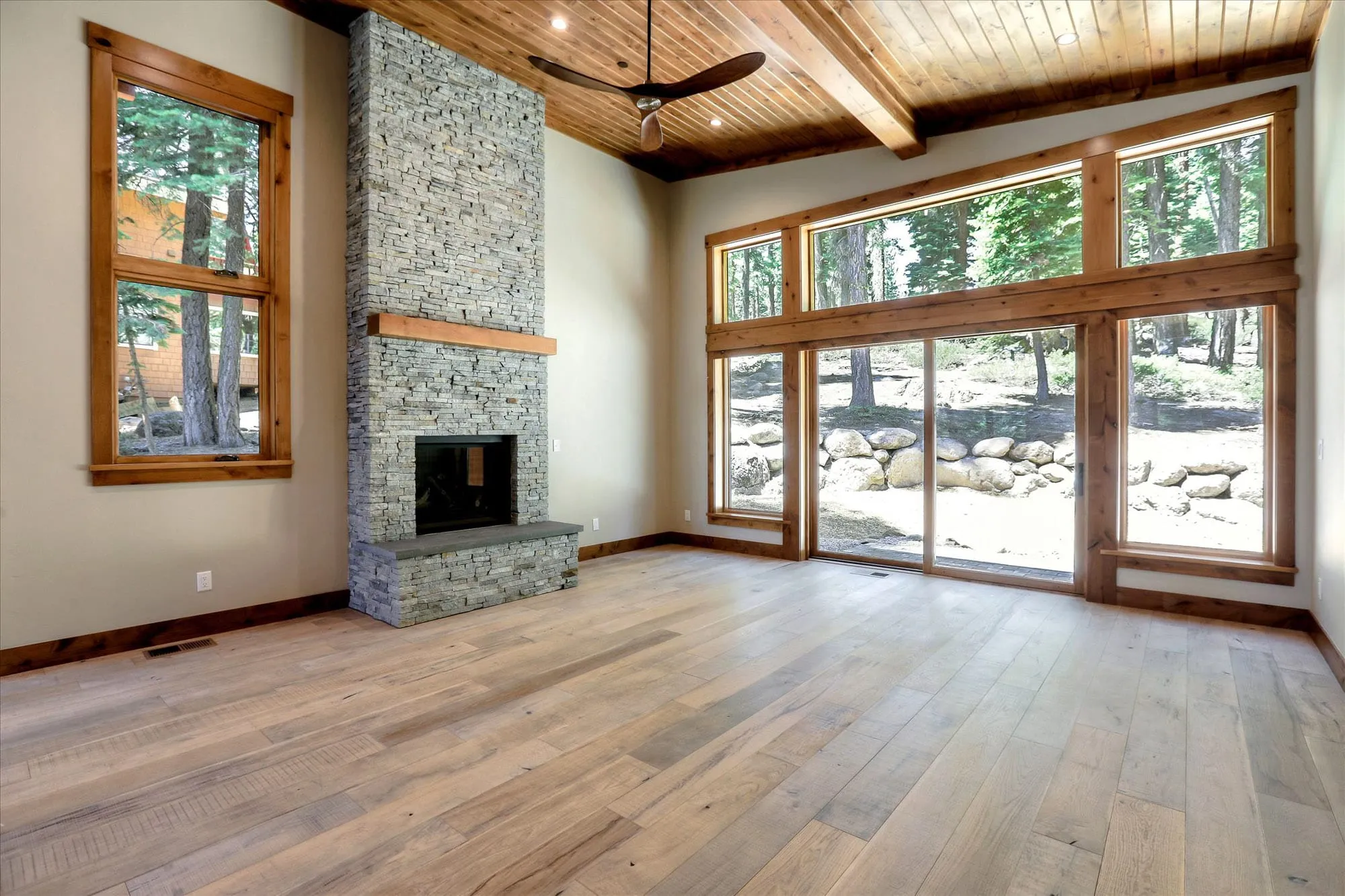 Interior living room with tall stacked-stone fireplace, wood plank ceiling, wide-plank floors and large glass doors