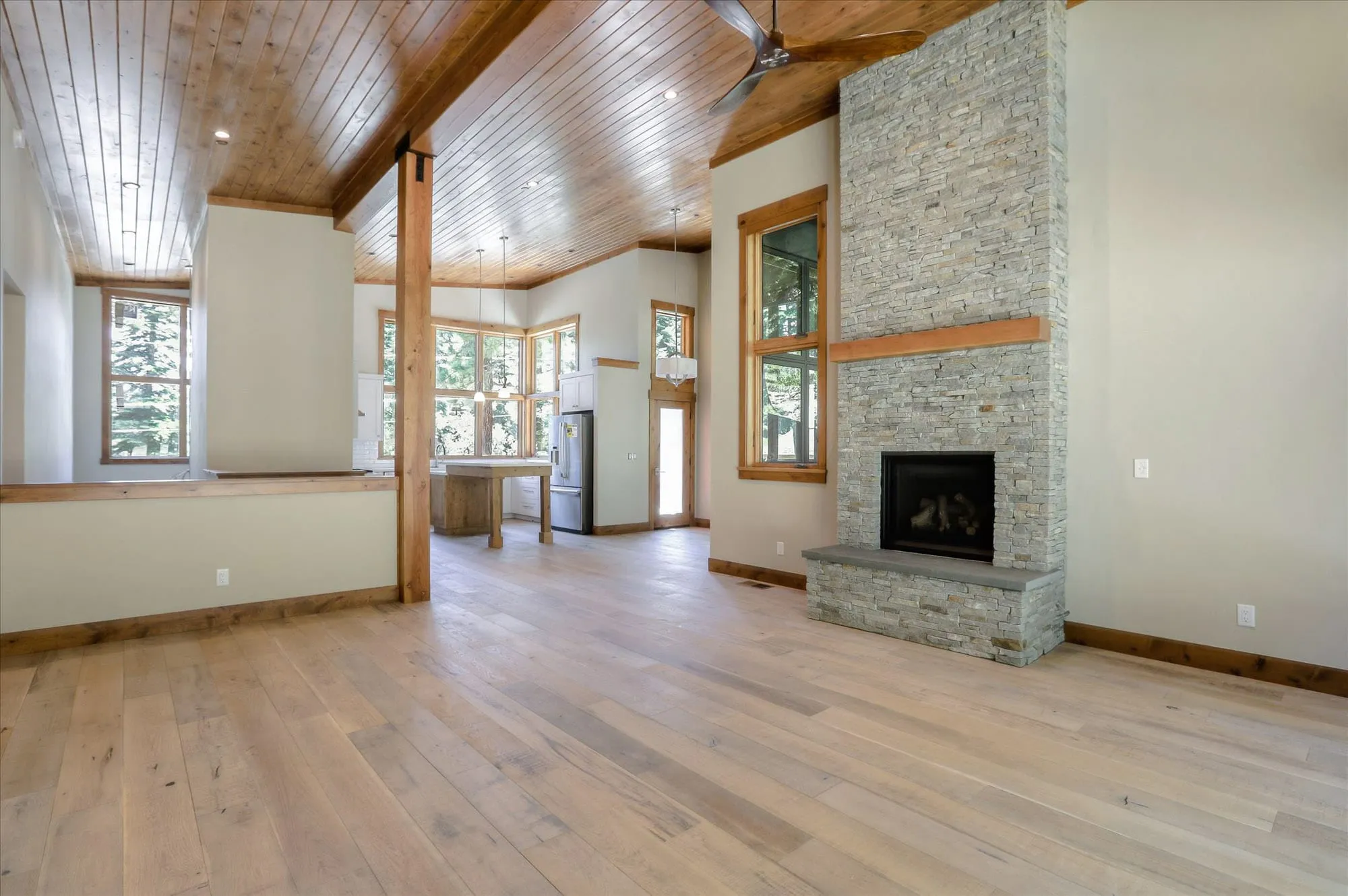 Open living area with stacked-stone fireplace, wood plank ceiling and timber post, leading to the kitchen beyond