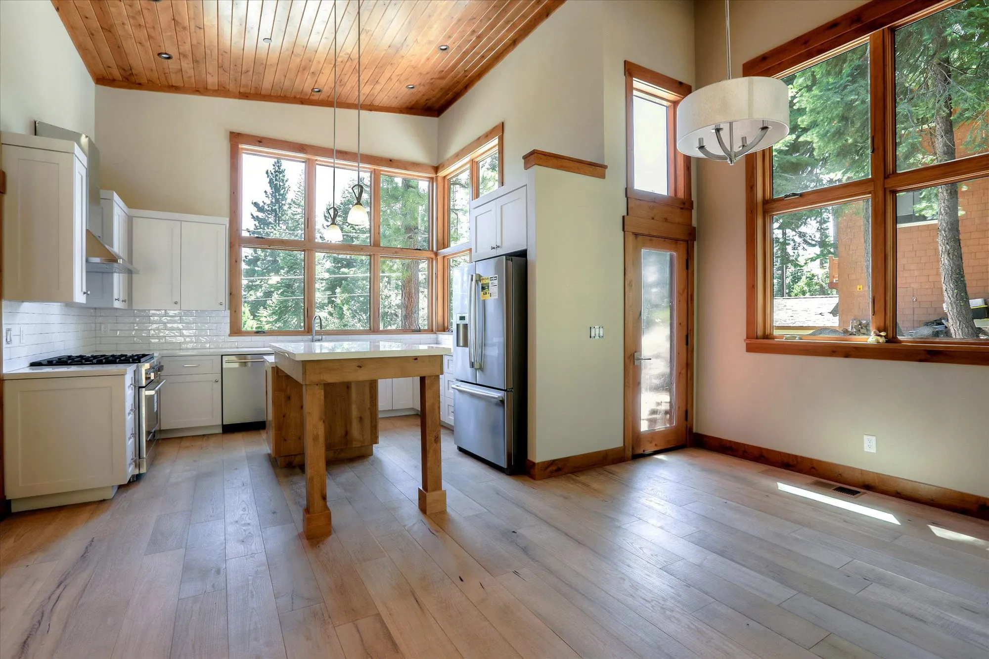 Kitchen with white cabinets, central wood island, stainless appliances, wood ceiling and tall windows onto trees
