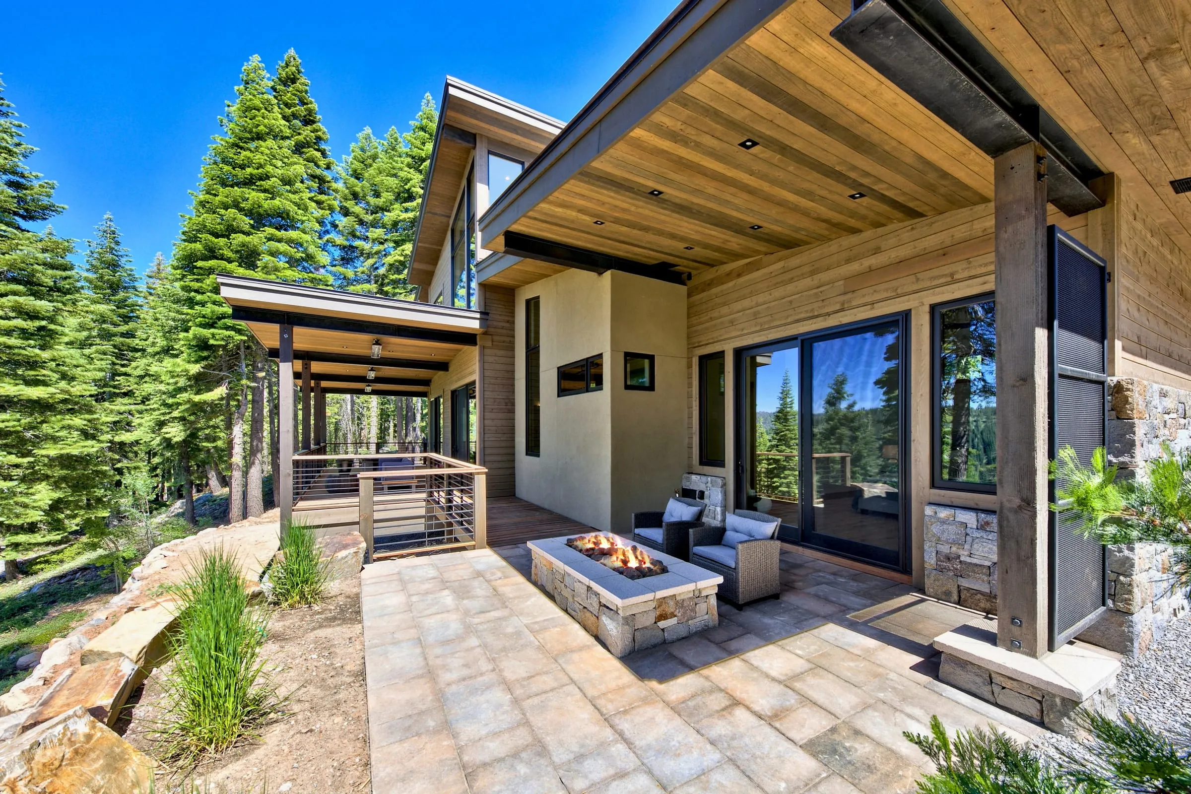 Stone-paved terrace with a fire table under a deep wood-ceilinged roof, beside a covered deck among tall firs