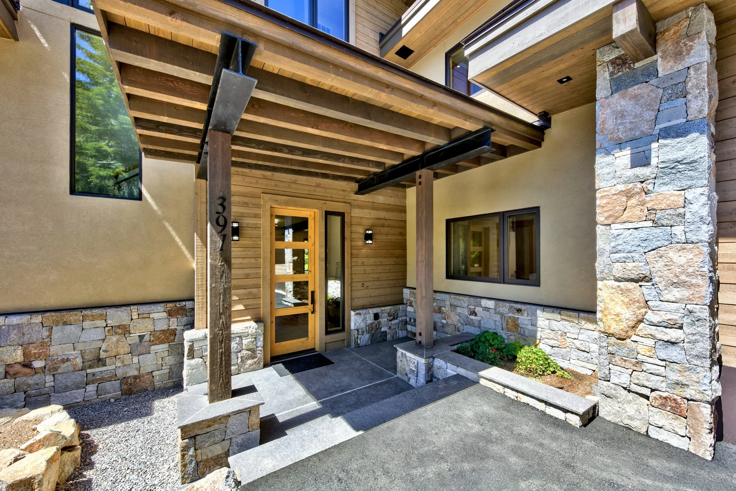 Entry porch with timber posts and steel beams, a glass-paneled wood door, stucco walls and a tall stacked stone pier