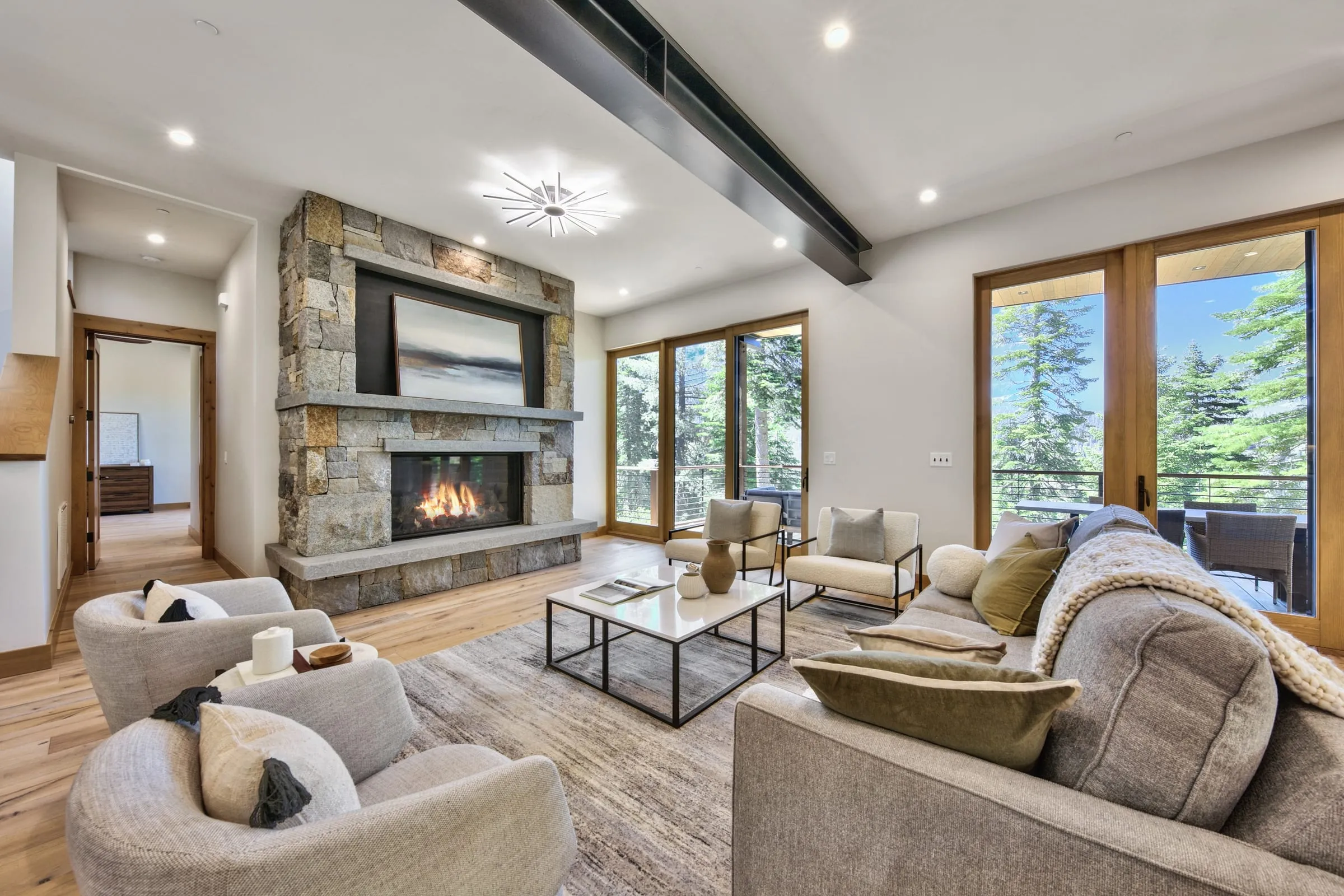 Living room with a stacked stone fireplace, a steel ceiling beam, light upholstered seating and glass doors to a deck among the trees