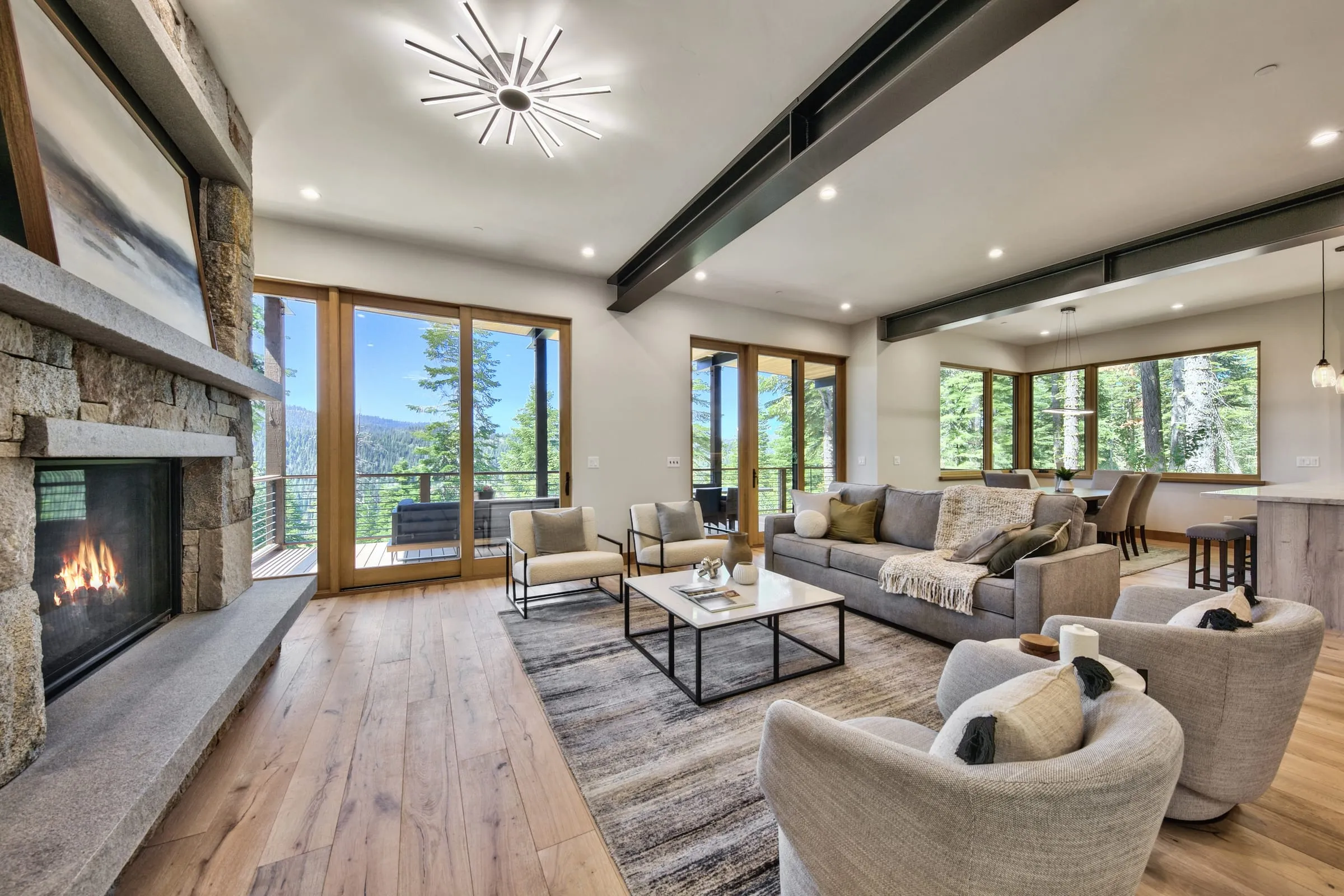 Living room with a stone fireplace, wide-plank oak floors and corner windows and glass doors to a deck with a forest view