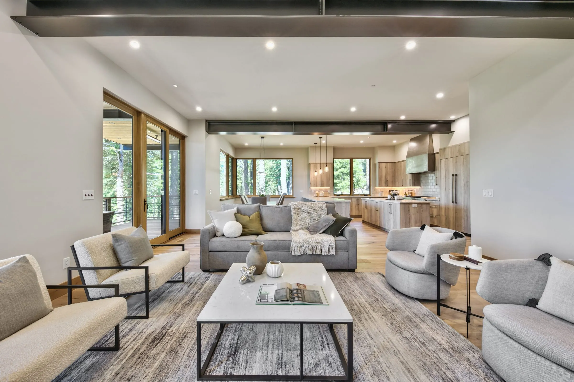 Open living room under a steel beam, looking toward the dining area and kitchen, with glass doors to a deck on the left