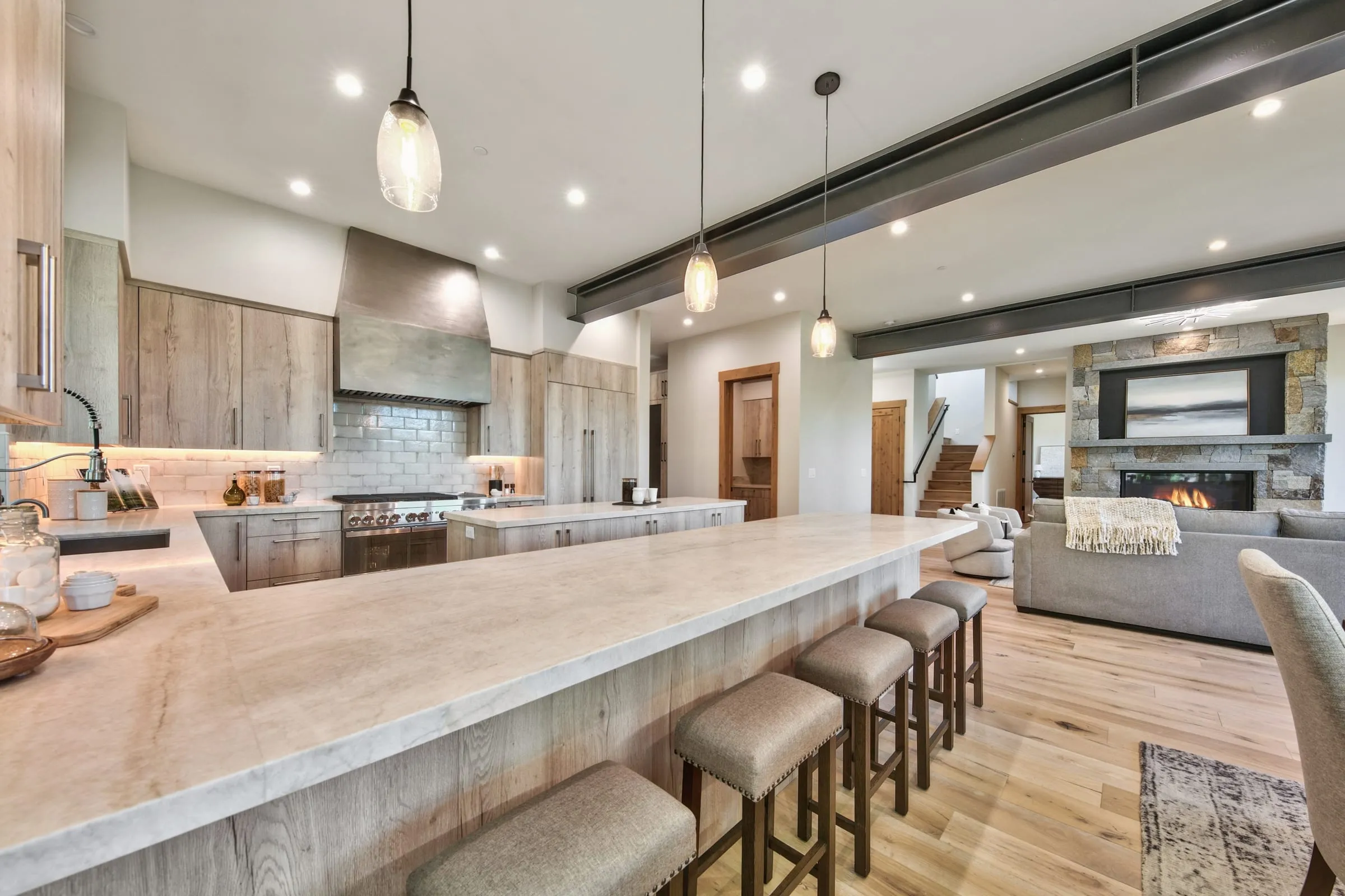 Kitchen with a long stone-topped island and stools, light wood cabinets, a metal hood and the living room fireplace beyond