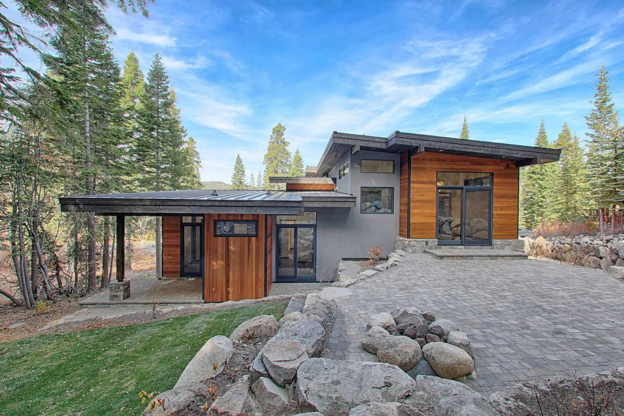 Modern mountain home with cedar siding, stone walls, shed roofs and a wraparound deck with cable railing among tall pines