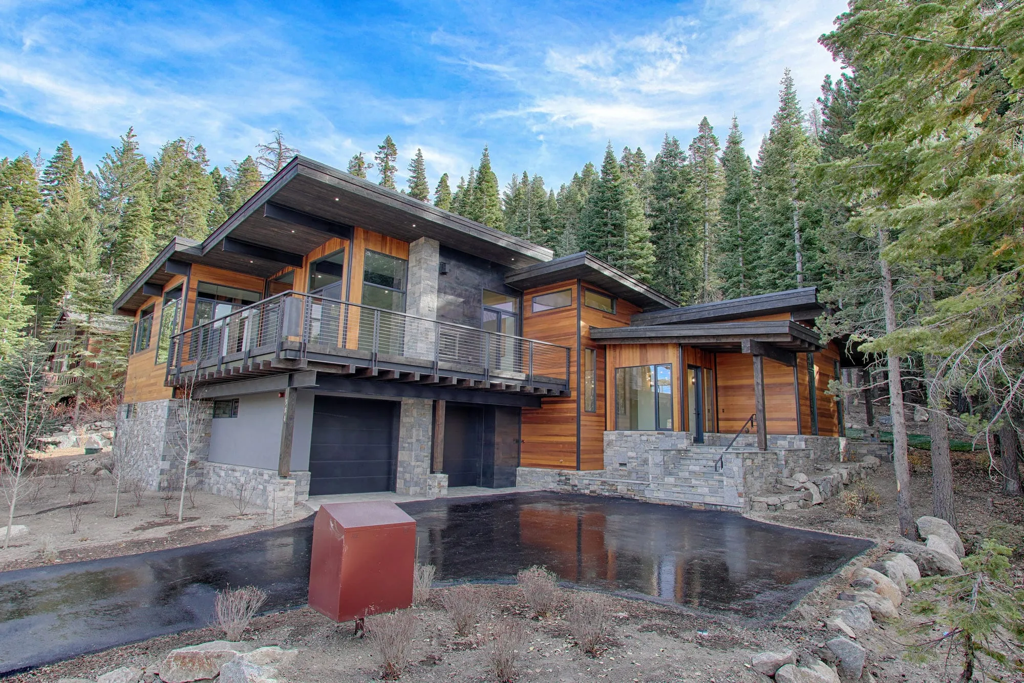 Front exterior of a modern home with cedar and gray siding, stone piers, dark garage doors and a cable-rail deck above