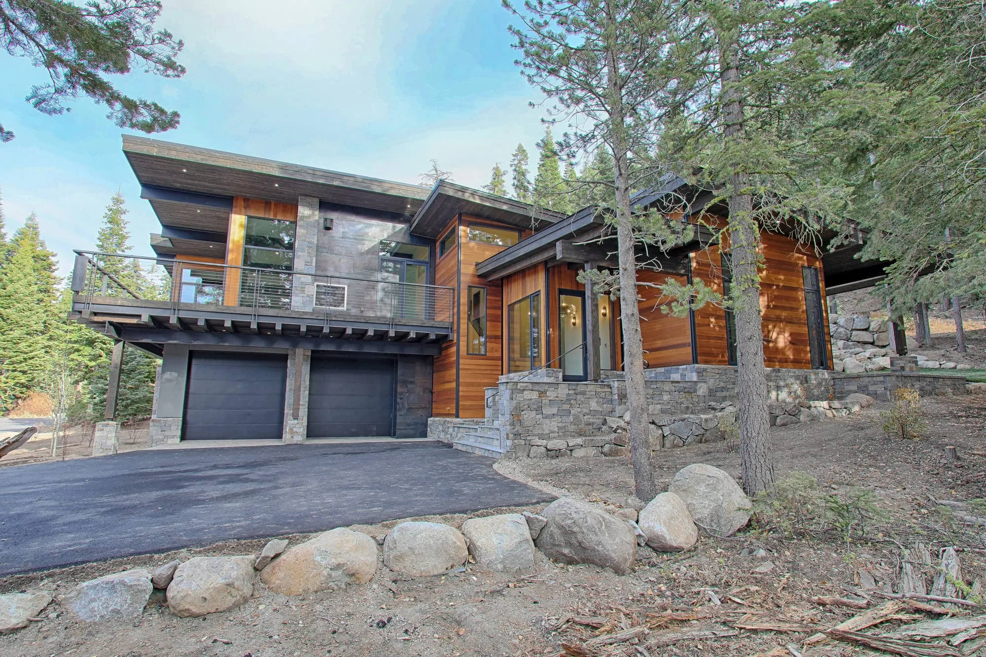 Corner view of a modern wood-and-stone home with two dark garage doors, upper deck and boulders along the driveway