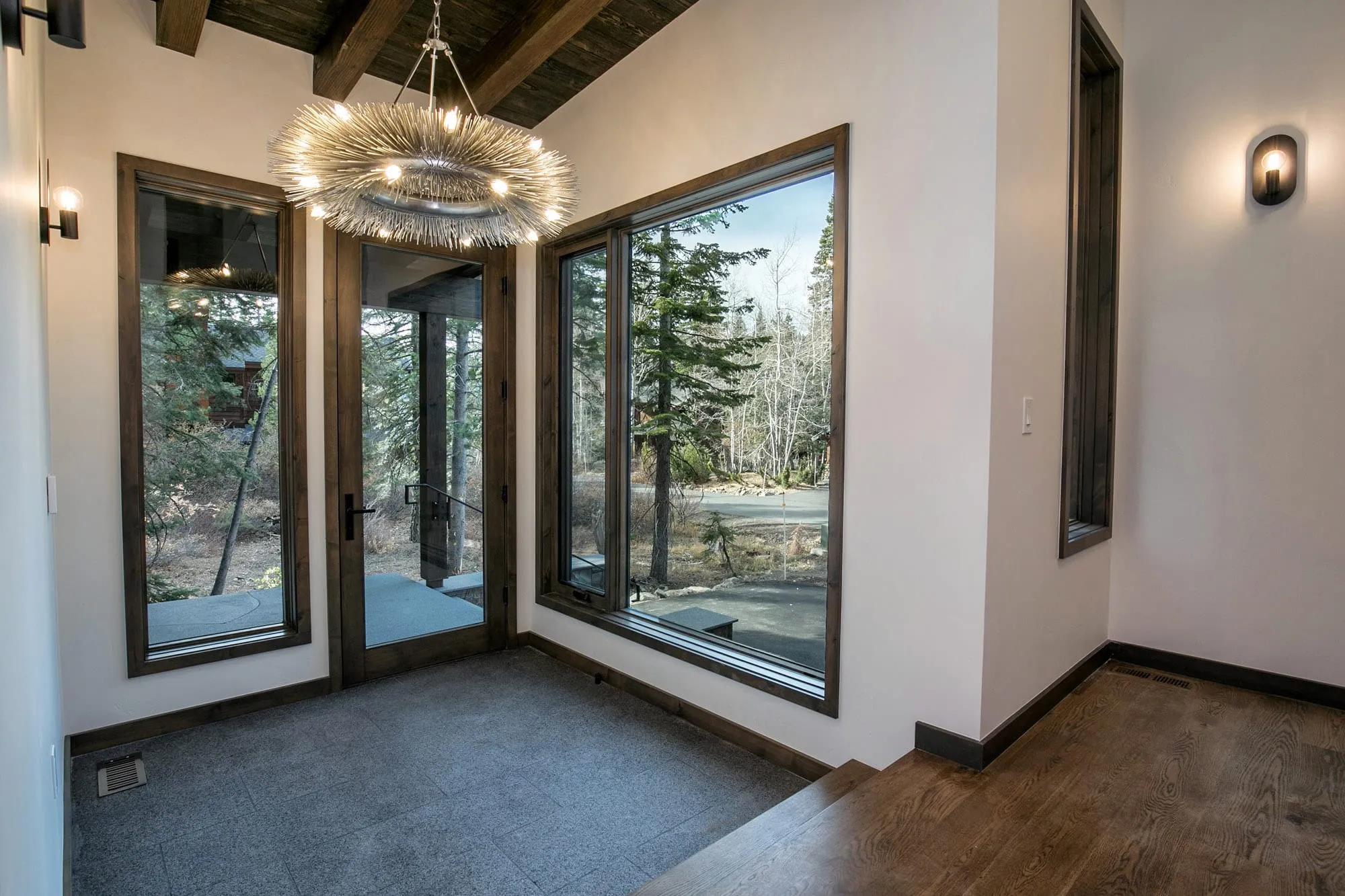 Entry foyer with a burst-style chandelier, stone floor, glass door, tall window onto pines and a few steps up to wood floor