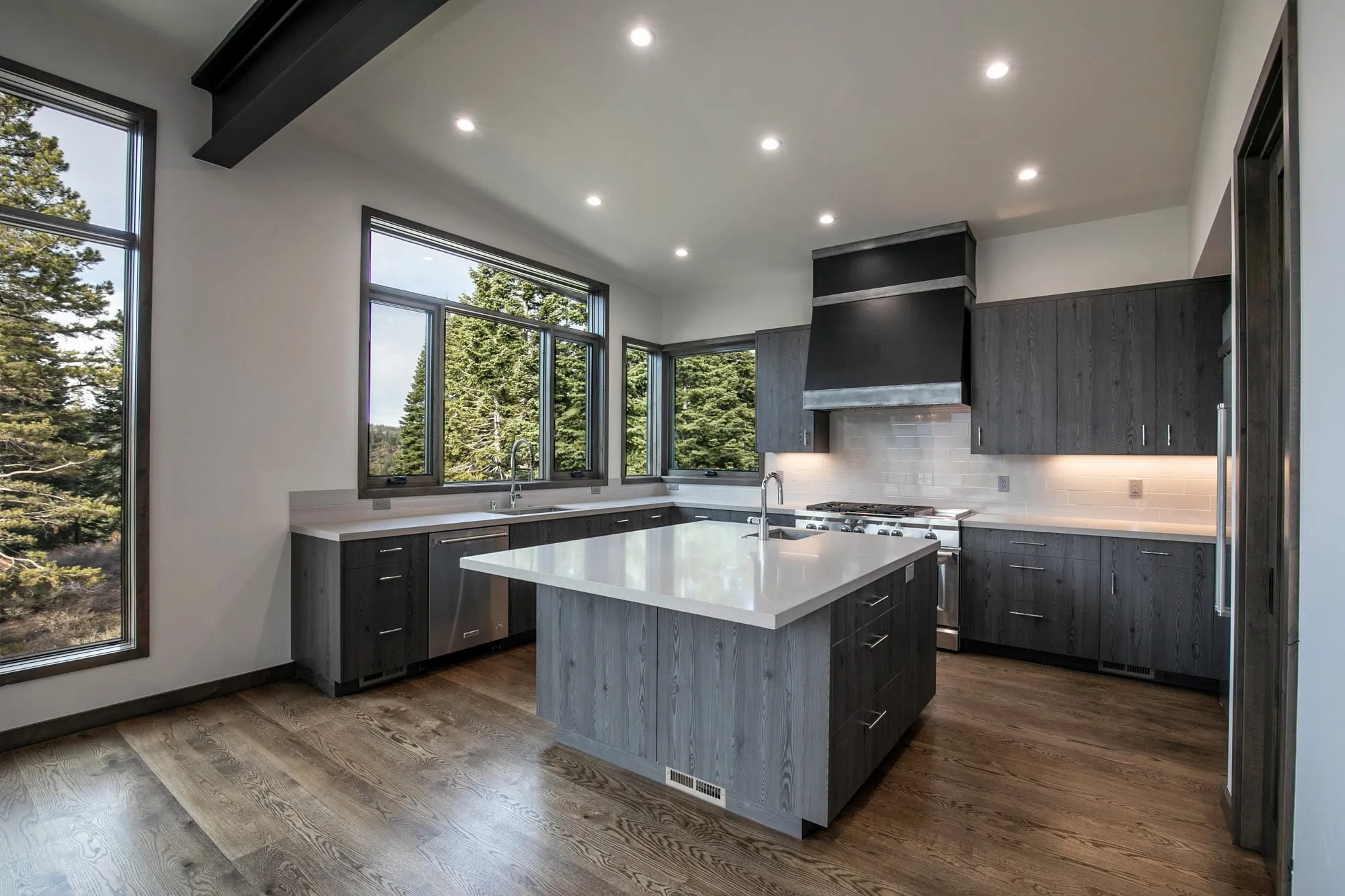 Kitchen with gray wood-grain cabinets, white quartz island, black range hood and windows onto pine trees