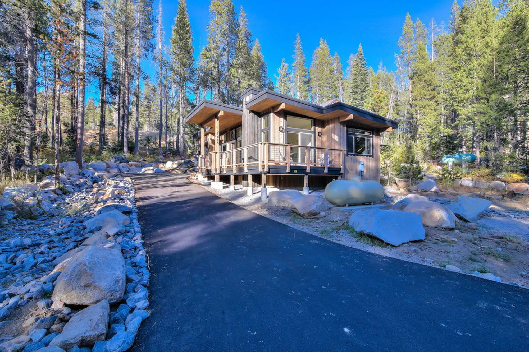 Small shed-roof cabin with a covered deck seen from a paved drive, surrounded by boulders and tall pines