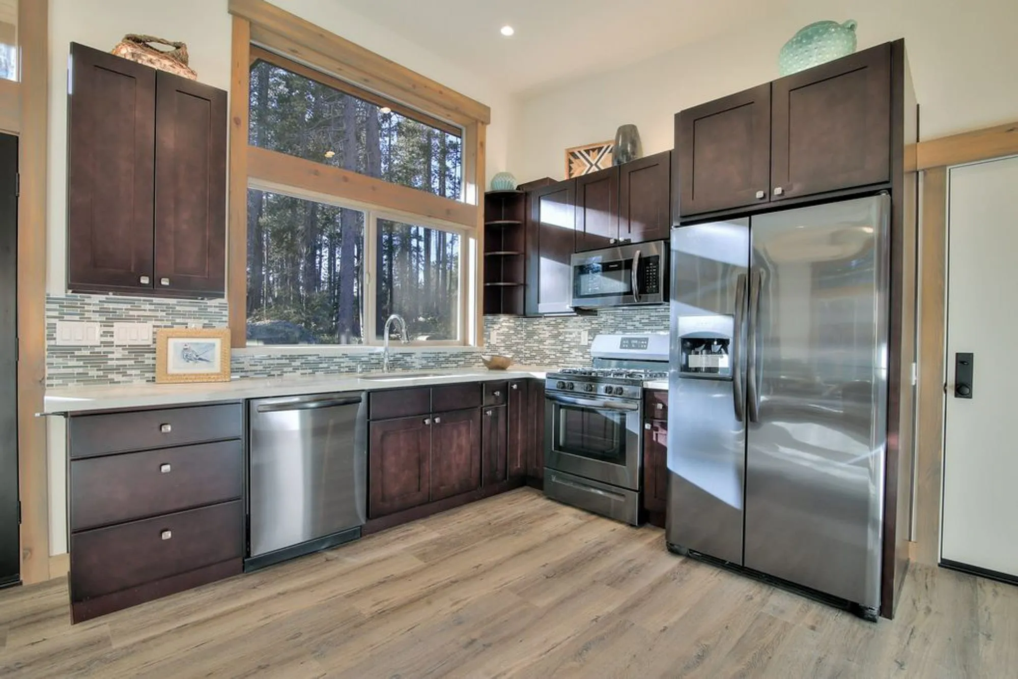 Compact kitchen with dark cabinets, glass tile backsplash, stainless appliances and a window looking out to pine trees